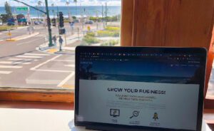 A laptop displaying a business website is positioned on a desk near a window, with a clear view of a street and the ocean in the background.
