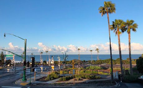 Intersection beside a coastal area with palm trees, a clear blue sky, and the ocean visible in the background. Traffic lights and street signs can also be seen.