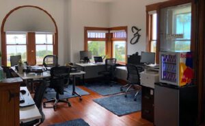 An office space with multiple desks and chairs, computer monitors, a Red Bull fridge, and large windows letting in natural light.