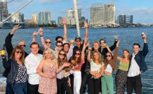 Group of people smiling, holding drinks, and posing with arms raised on a boat, with a city skyline and waterfront in the background.