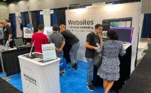 A trade show booth featuring a company's banner and a desk with promotional materials and multiple people engaging in conversations.