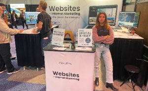 A woman stands at a booth labeled, Websites + Internet Marketing, with promotional signs for services at a trade show. People are interacting in the background.