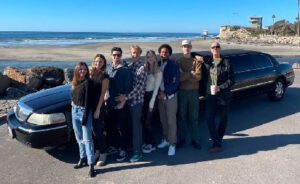 A group of eight people stand together in front of a black limousine by the beach. The ocean and a clear blue sky are visible in the background.