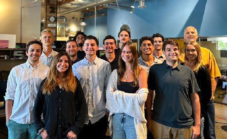 A group of 14 people stand closely together, posing for a photo in an indoor setting with a high ceiling and an industrial design.