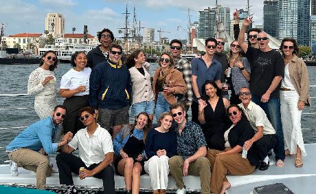A group of people poses together on a boat with a cityscape and sailing ships in the background.