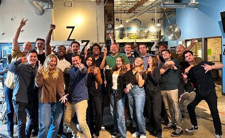 A group of people posing enthusiastically in an indoor setting, with some raising their arms and making playful gestures. Industrial-style interior with metal ducts and large windows.