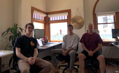 Three people are sitting in an office with wooden windows and a view of palm trees and the ocean. Computer desks and office equipment are visible in the background.