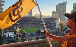 A person waves a San Diego Padres flag on a balcony overlooking a crowded baseball stadium with city buildings in the background.