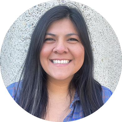 A person with long dark hair smiles while wearing a blue plaid shirt, standing in front of a textured concrete wall.