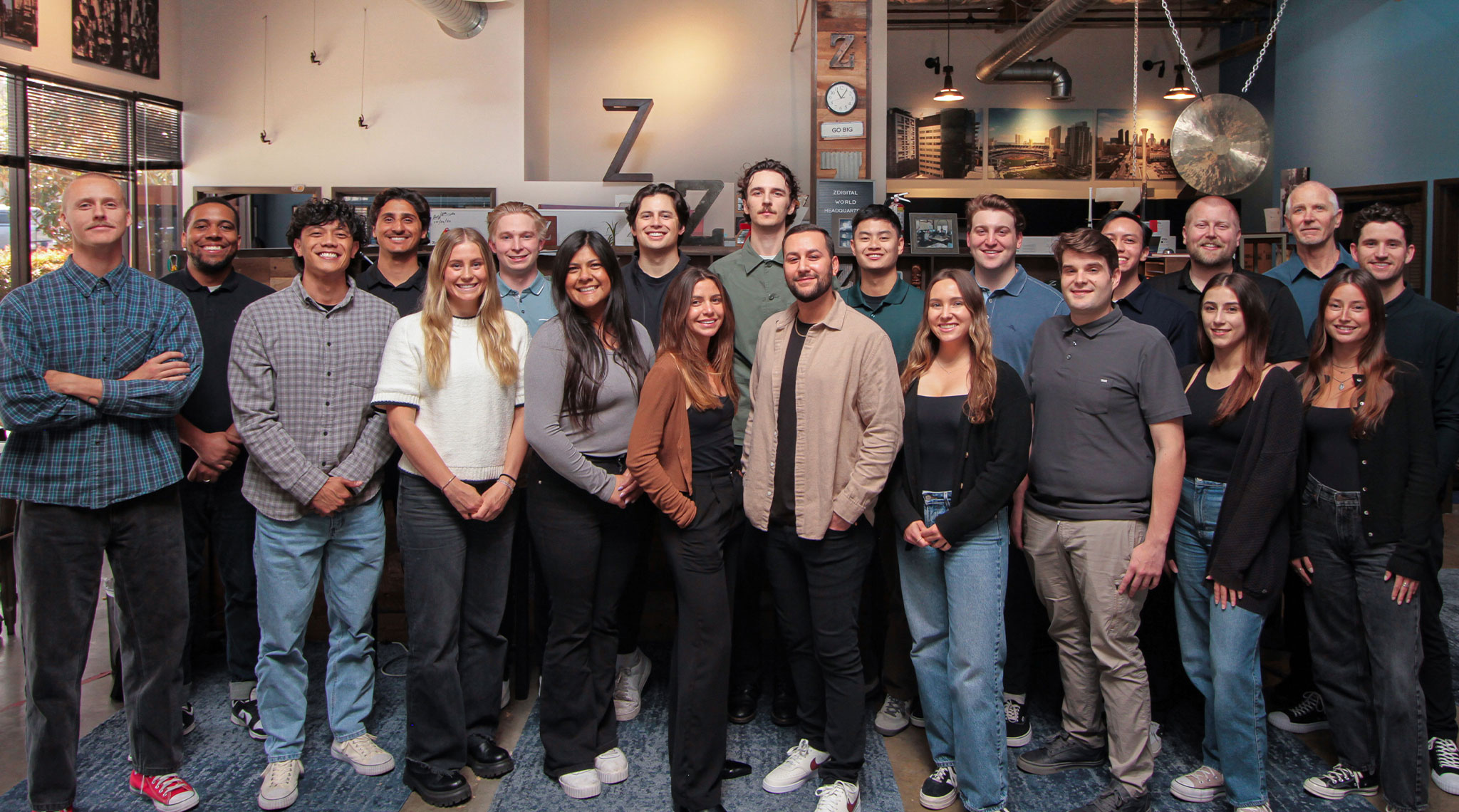 home-zdigital-hero-4-2-25 A diverse group of people standing indoors, posing for a group photo in casual attire. Some are smiling, and a large gong is visible in the background.