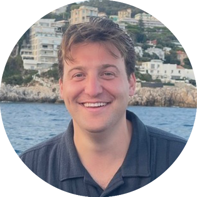 A man with short brown hair smiles at the camera while standing in front of water, with buildings and greenery visible in the background.